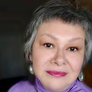 A 56 year old woman looking directly into the camera wearing a lavender turtleneck and wampum necklace.